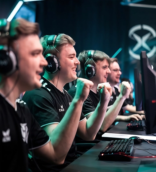 Vier esports-spelers met een headset zitten achter computerschermen en vieren hun overwinning met opgeheven vuisten. De ruimte wordt verlicht door neonlichten, wat zorgt voor een energieke sfeer