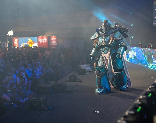 Een persoon in een gedetailleerd, futuristisch blauw harnas loopt over het podium tijdens een bruisend esports-festival, terwijl het publiek toekijkt en rookeffecten voor een meeslepende sfeer zorgen