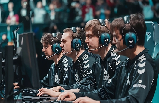 Vijf geconcentreerde esports-spelers in bijpassende uniformen en met headsets op, zittend achter gamingcomputers in een arena, met een wazig publiek op de achtergrond