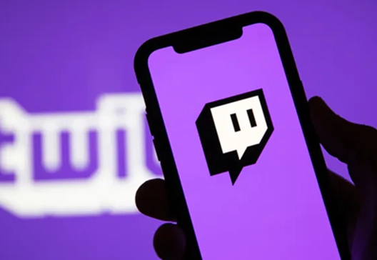Een hand houdt een smartphone vast met het Twitch-logo op het scherm, tegen een wazige paarse achtergrond waarop een groter Twitch-logo te zien is